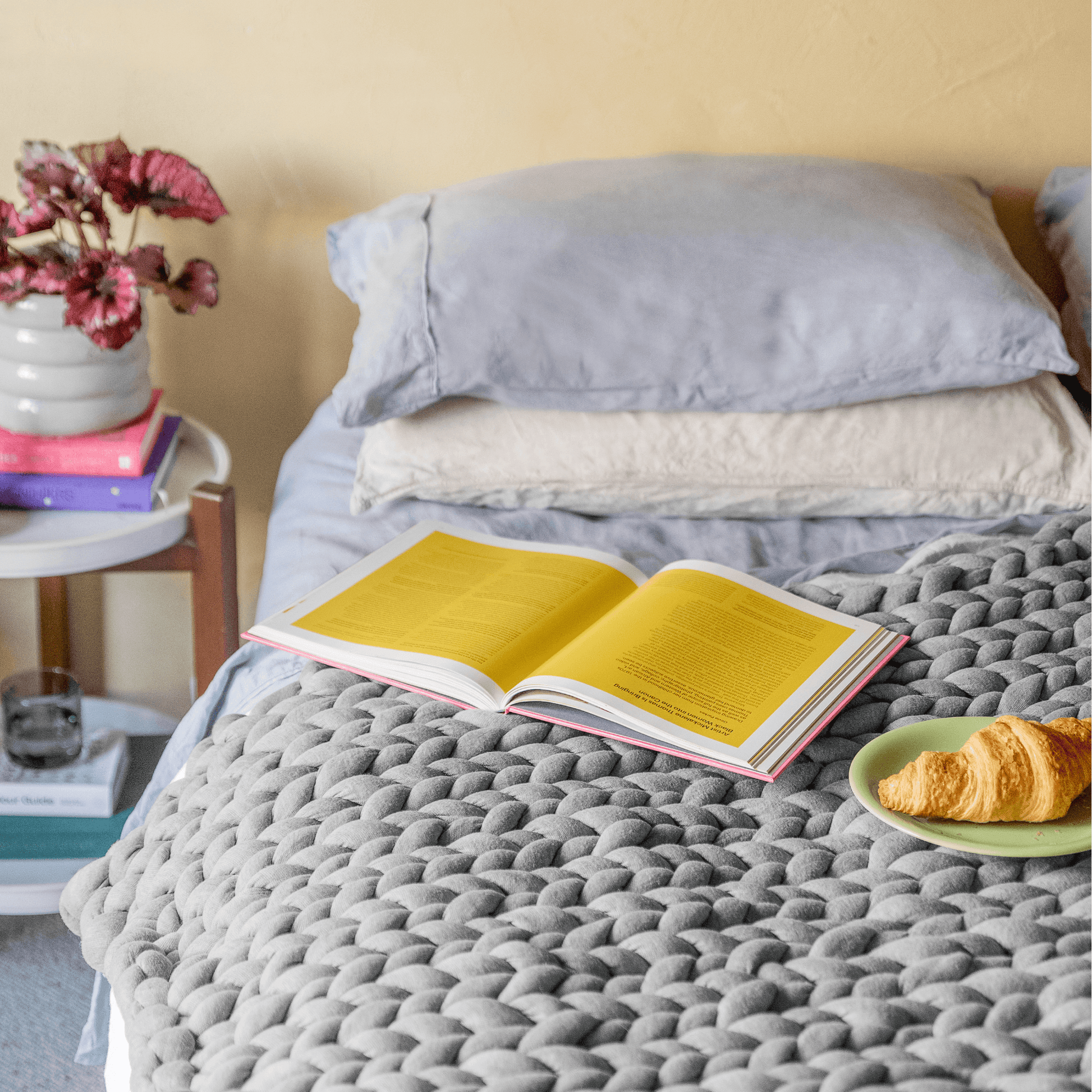 Finished grey chunky knit throw on top of a bed with a book and croissant